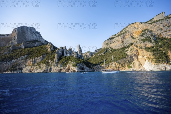 Excursion boat off picturesque rocky coast, cliffs with caves in morning light, L'Aguglia rock pin, blue sea and Cala Goloritzé beach, Golfo di Orosei, Baunei, Sardinia, Italy