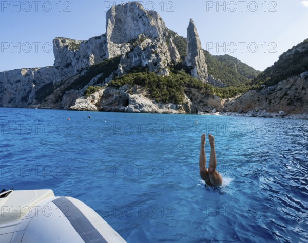 Young woman jumping from a boat into the water, picturesque rocky coast, cliffs with L'Aguglia rock pin, blue sea and Cala Goloritzé beach, Golfo di Orosei, Baunei, Sardinia, Italy