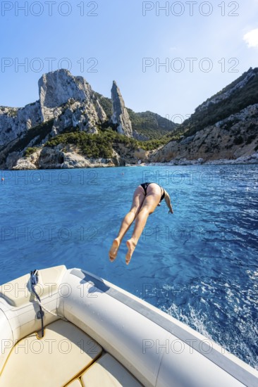 Young woman jumping from a boat into the water, picturesque rocky coast, cliffs with L'Aguglia rock pin, blue sea and Cala Goloritzé beach, Golfo di Orosei, Baunei, Sardinia, Italy