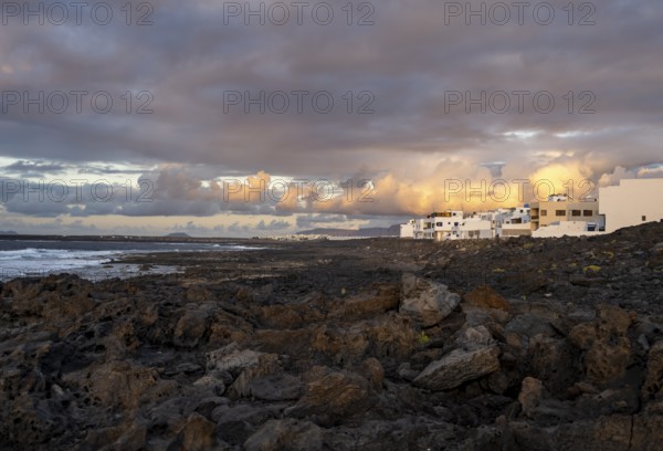 Black coast of volcanic rocks behind typical white houses of the village of La Santa, at sunset, Lanzarote, Canary Islands, Spain
