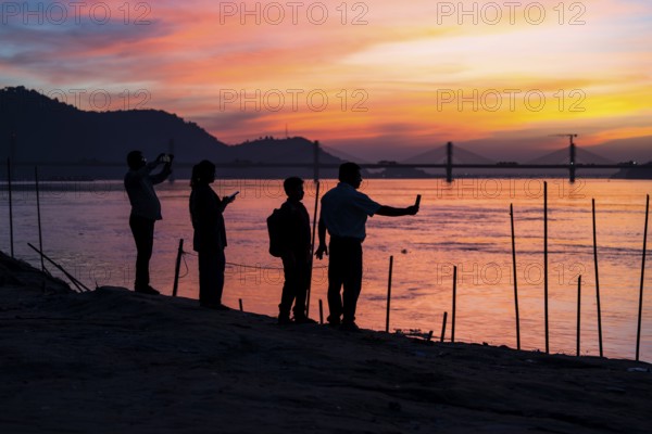 People enjoy the vibrant sunset by the Brahmaputra riverside, capturing photos and moments against the scenic backdrop of a bridge and glowing sky, on November 4, 2025 in Guwahati, India
