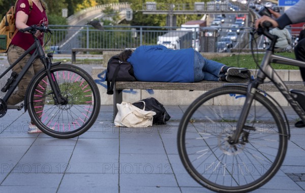 Man sleeping on a bench, travelling bag serves as pillow, in the city centre, passers-by walk by