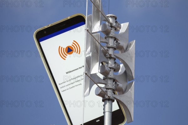 (Photomontage) Close-up of a siren, next to it the open warning app NINA on a smartphone