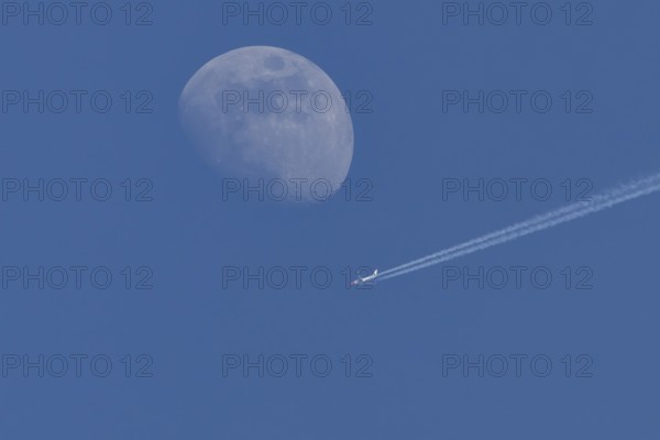 Boeing 737 jet passenger aircraft of Norweign Air with a vapour trial or contrail behind flying in a blue sky passing by the moon, England, United Kingdom