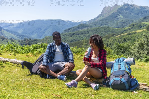 Two hikers are taking a break during their mountain trek, sitting on a log, drinking water and enjoying the scenic view