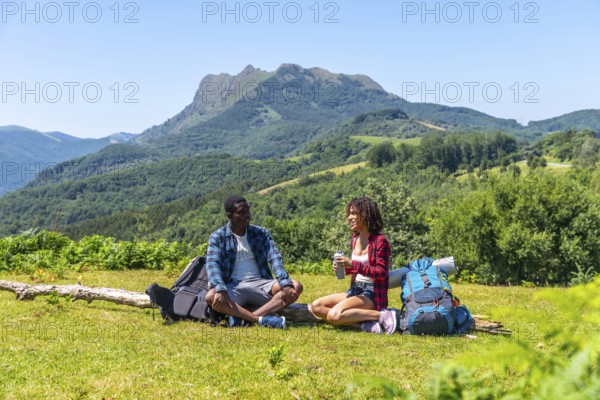 Two hikers are taking a break, sitting on grassy mountaintop and enjoying refreshing drinks with backpacks beside them