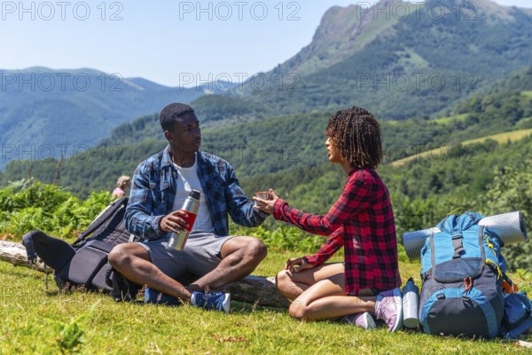 Young hikers sharing a hot drink from a thermos while resting on a mountain top during a summer hike