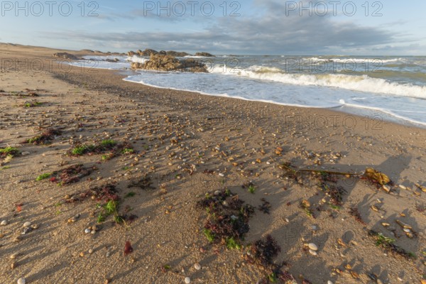 The waves crash gently against a rocky shoreline at sunset, creating a tranquil atmosphere. The sandy beach is strewn with pebbles and surrounded by a beautiful sky. Plage de l'Aubraie, Les Sables d'Olonne, Vendee, France