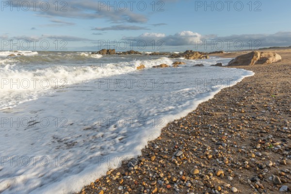 A wide sandy beach stretches out with scattered pebbles and beach shells. Gentle waves crash against the shore under a partially cloudy sky during late afternoon. Plage de l'Aubraie, Les Sables d'Olonne, Vendee, France
