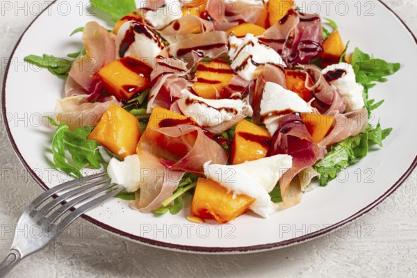 Salad with slices of ripe melon, prosciutto, mozzarella and fresh arugula, decorated with balsamic glaze
