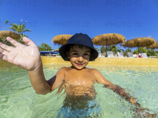 Smiling child in a sun hat waves while playing in a clear swimming pool, surrounded by straw umbrellas and tropical palm trees