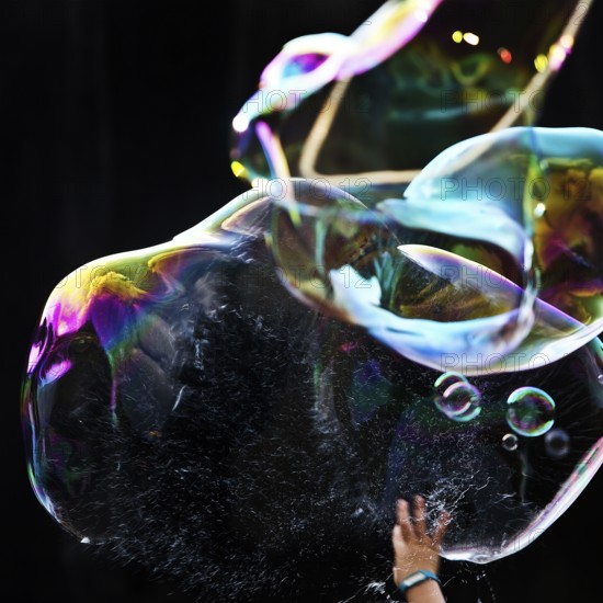 A hand bursts a large iridescent soap bubble against a black background, Recklinghausen, Germany