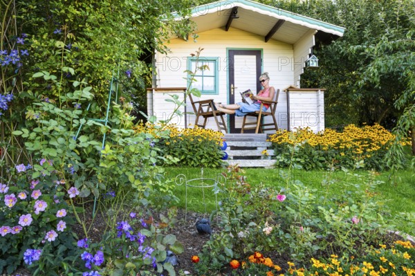 A woman sits relaxed on the small veranda of a garden shed and reads. Blooming summer flowers in the garden. Germany