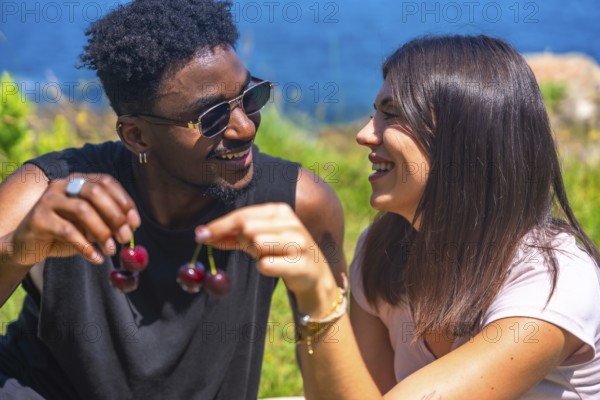 Young man and woman laughing together while sharing fresh cherries, savoring the joy of a sunny summer day in a beautiful outdoor setting