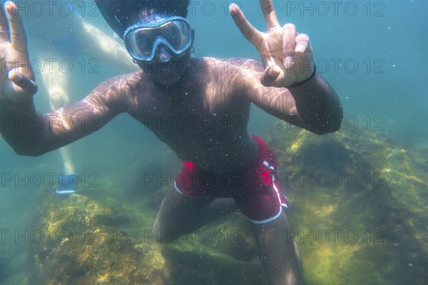 Young man snorkeling underwater, flashing the peace sign with both hands, enjoying the vibrant blue sea and colorful coral reef