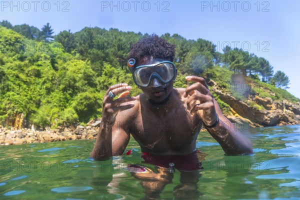 Young man wearing a snorkel mask and gesturing while swimming in green, translucent water near a rocky, tree lined coast