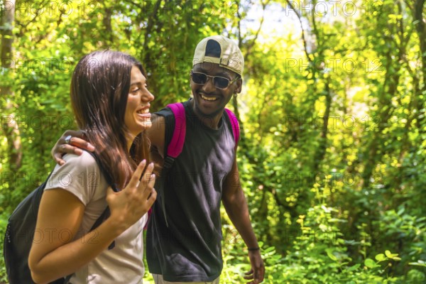 Happy multi ethnic couple enjoying trekking in a green forest, wearing backpacks and casual clothes