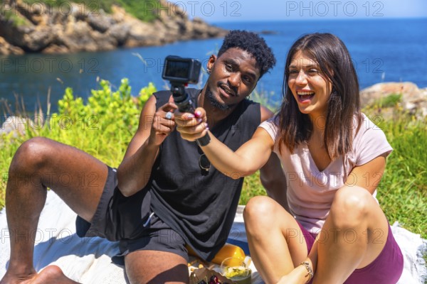 Happy young multi ethnic couple filming with action camera while having picnic by the sea during summer vacation