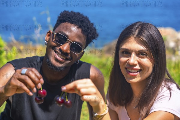 Interracial couple smiling while sharing fresh cherries, enjoying a sunny summer vacation by the sea, radiating joy and togetherness