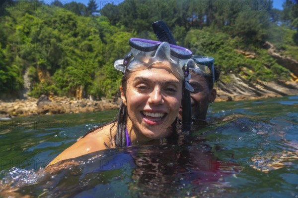 Interracial couple snorkeling together in the ocean, surrounded by a vibrant green coastline, enjoying a joyful summer adventure