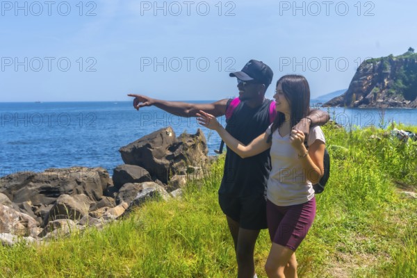 Interracial couple hiking along a stunning coastline, joyfully pointing at a breathtaking view in the distance, sharing a moment of adventure