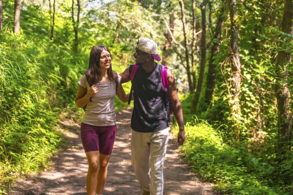 Interracial couple enjoying a hike on a forest path, wearing backpacks and casual clothes, engaging in conversation
