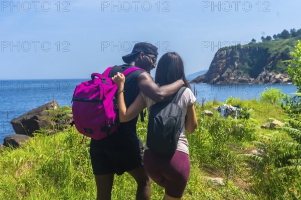 Interracial couple with backpacks walking on a path by the sea, embracing and enjoying the scenic view during a sunny summer day