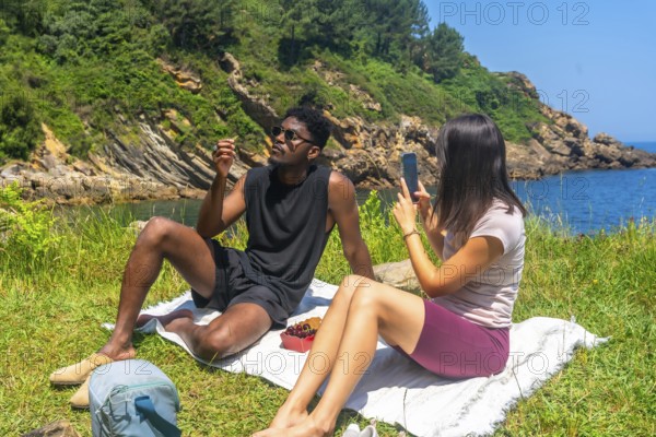 Young woman takes a photo of her friend during a scenic picnic by the sea, capturing memories of their travel adventure