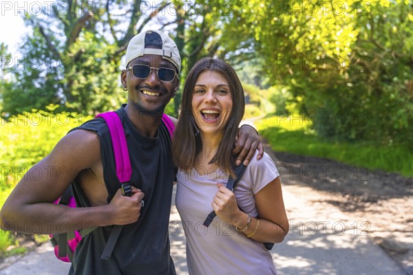 Interracial couple hiking on a sunny summer day, embracing each other and sharing joyful smiles amidst the vibrant green trees