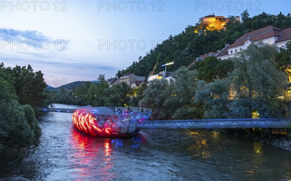 Night-time view of the Murinsel floating island on the river Mur with Schlossberg castle hill in the background, Graz, Austria