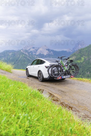 White car with bicycles on mountainous road, surrounded by green landscape and cloudy sky, electric car on holiday, Dolomites, South Tyrol, Dolomites, Italy