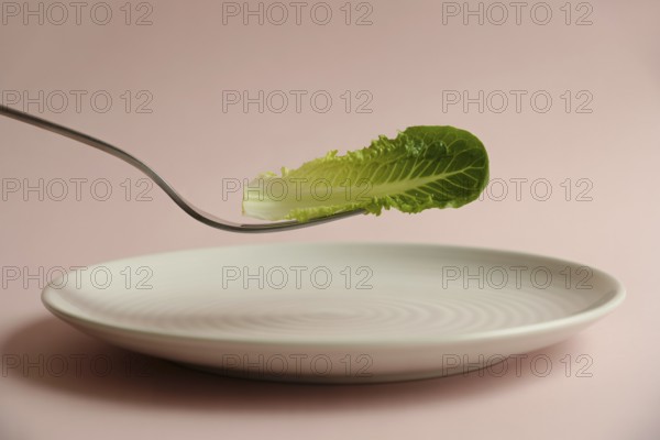Minimalist close-up of fork holding lettuce leaf above white plate on pink background. Concept of strict diet and unhealthy eating, AI generated