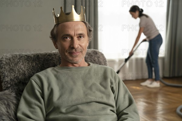 Portrait of middle-aged man with golden crown relaxing on couch while woman vacuums in background. Concept of patriarchal carework imbalance and male privilege. Generative ai, AI generated