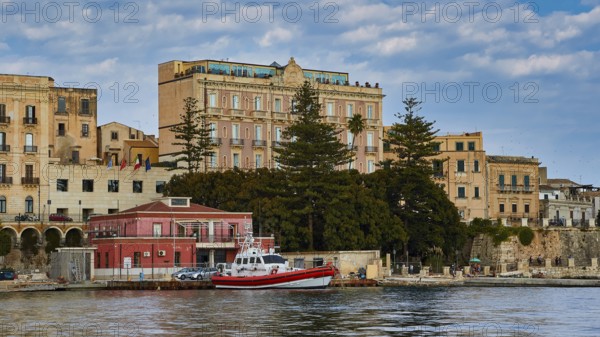 Boats in front of historic buildings and trees on a ponderous coast under a cloudy sky, boat trip around the island of Ortigia, Isola di Ortigia, Syracuse, Sicily, Italy