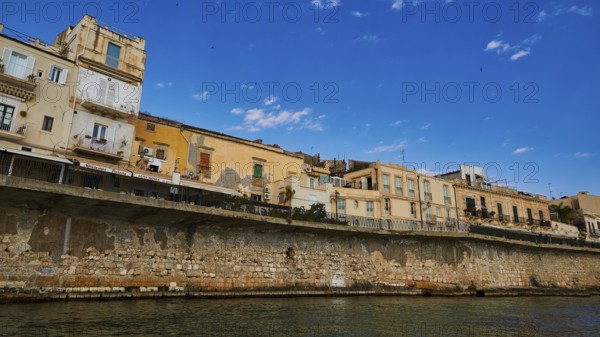 Row of historic houses on a stone seawall under a clear sky, boat trip around the island of Ortigia, Isola di Ortigia, Syracuse, Sicily, Italy