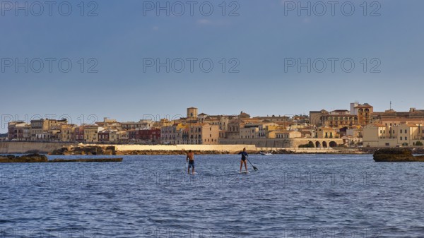Two paddleboarders on the sea in front of a town with historical buildings under a blue sky, boat trip around the island of Ortigia, Isola di Ortigia, Syracuse, Sicily, Italy