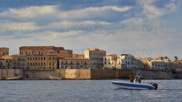 Boat with two people in front of historic coastline and cloudy sky, boat trip around the island of Ortigia, Isola di Ortigia, Syracuse, Sicily, Italy