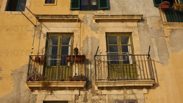 Weathered historic building façade with two balconies, windows and flowers under sunlight, Ortigia Island, Isola di Ortigia, Historic Centre, Syracuse, Sicily, Italy