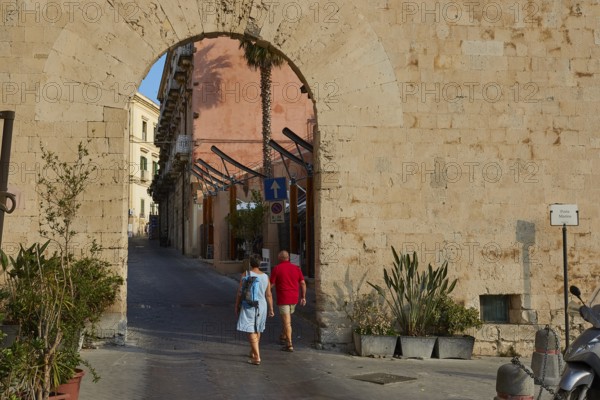 People crossing a historic stone city gate surrounded by plants, Ortigia Island, Isola di Ortigia, Historic city centre, Syracuse, Sicily, Italy
