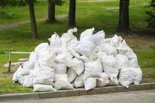 Multiple white heavy-duty garbage bags filled with waste material are piled up in a green park area. Trees surround the site under bright daylight, indicating a need for cleanup efforts