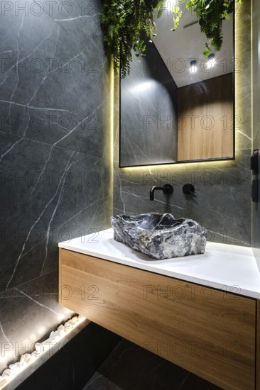A sleek bathroom with a striking stone sink mounted on a wooden cabinet. The walls are tiled with black marble tiles