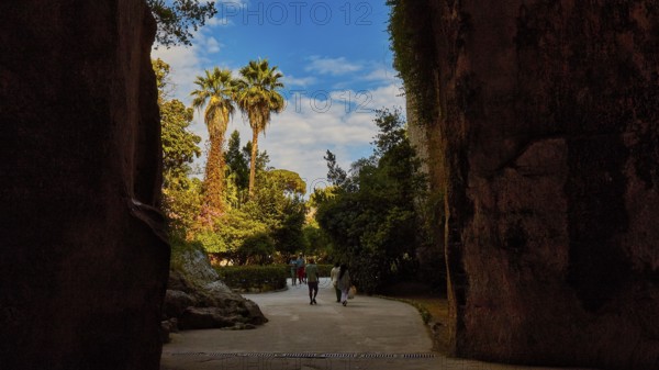 Cave Ear of Dionysus, Two people walking through a rocky passage with palm trees in the background in sunny weather, Archaeological Park Neapolis, Syracuse, Sicily, Italy