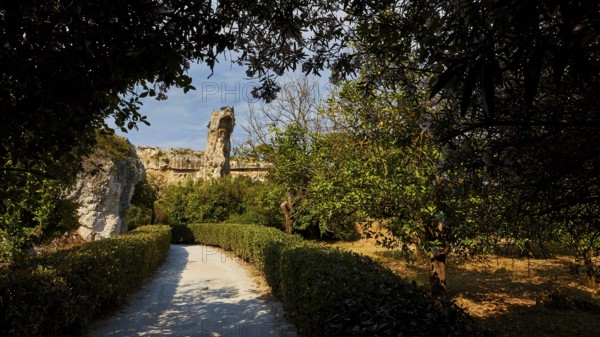 Park, A path leads through a sunlit landscape of ruins surrounded by trees and bushes, Neapolis Archaeological Park, Syracuse, Sicily, Italy