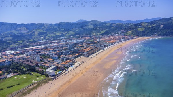 Aerial view of zarautz, gipuzkoa, showcasing the urban area meeting a golden sandy beach, with rolling green hills and the vast blue sea under a clear sky, Spain.