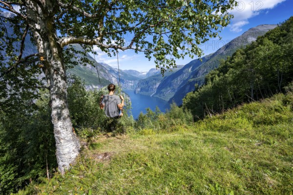 Young man swinging on a tree, view of idyllic fjord landscape of the Geirangerfjord at Blomberg Gård, near Geiranger, Møre og Romsdal, Norway