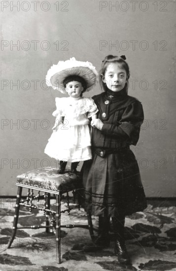 Girl doll dresses, 1930s, girl standing next to a chair carrying a doll in a dress, both in nostalgic clothes, historical photo