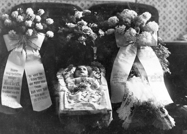Dead baby in a coffin, Infant mortality, 1920s, An infant lies in a coffin surrounded by floral wreaths and mourning ribbons, Historical photo