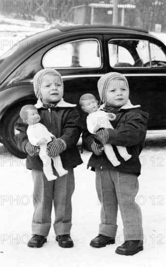 Twins, 1950s, Two children standing outside in the snow, holding dolls, car in the background, Historical photo