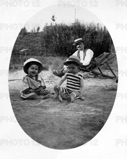 Father with 2 children beach deckchair, 1920s, Two children playing in the sand, an adult in the background, relaxed summer day, historical photo