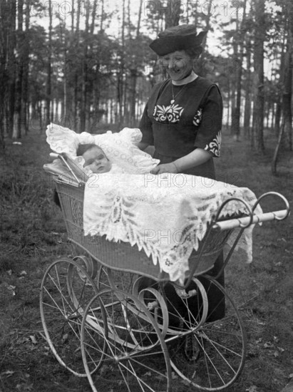 Couple with baby in pram, 1910s, woman in vintage dress next to a pram in the forest. A baby lies in the pram under an embroidered blanket, historical photo